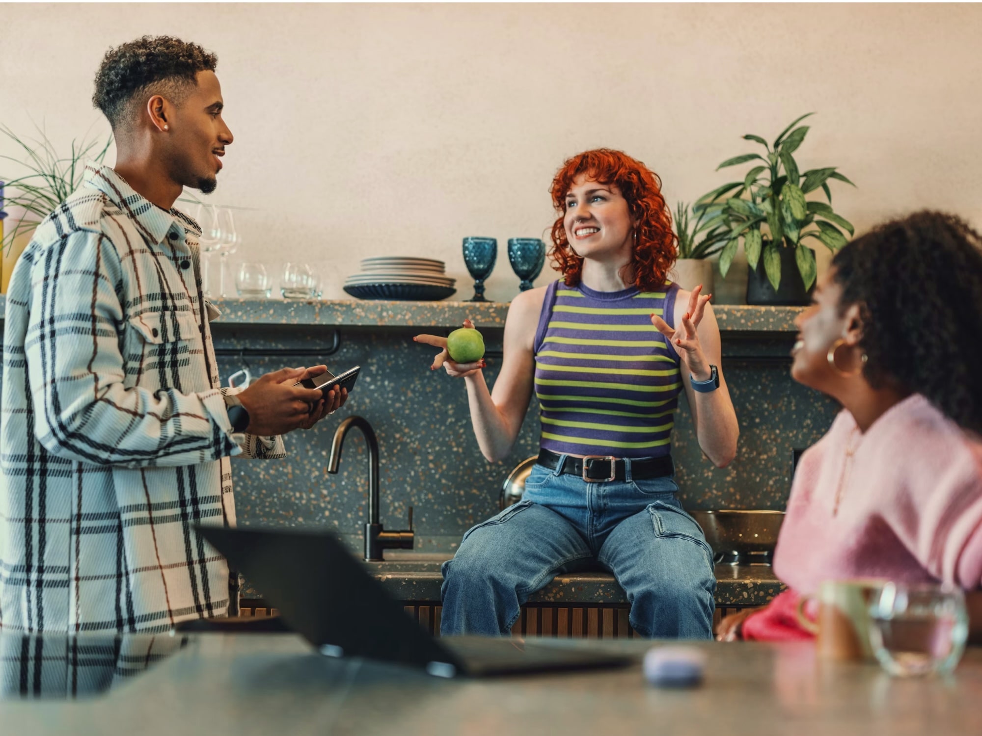 Three young adults having a lively conversation in a kitchen, one woman with red curly hair holding a green apple and smiling.