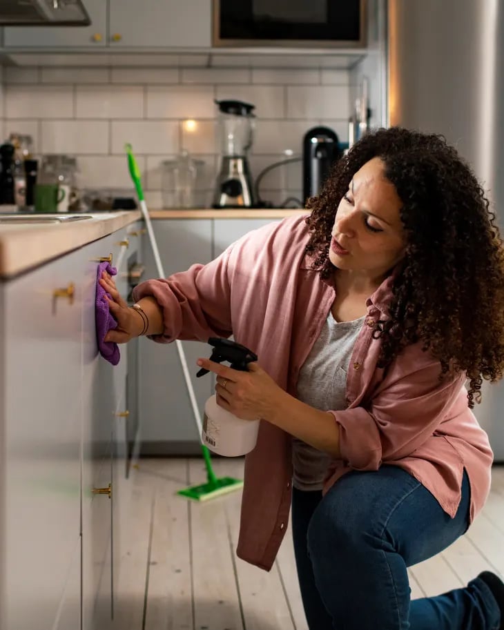 Woman with curly hair kneeling and cleaning kitchen cabinets with a purple cloth and spray bottle.