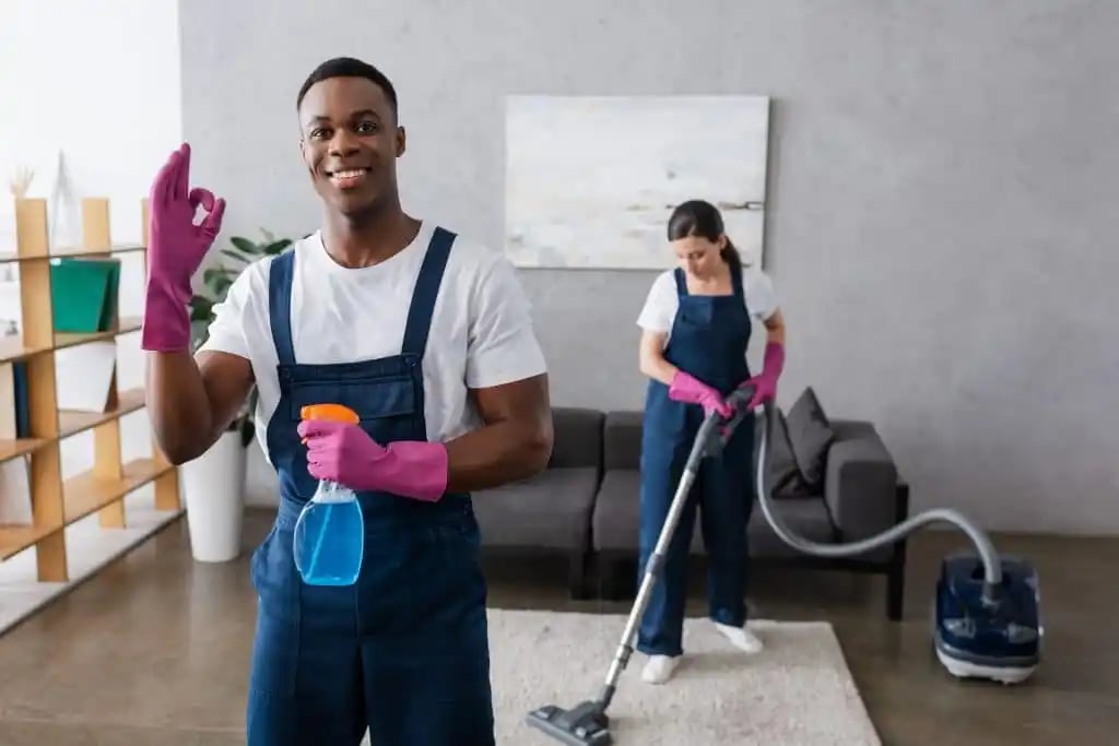 Two people wearing overalls and pink gloves cleaning a living room; one holding a spray bottle and making an OK gesture, the other vacuuming a rug.