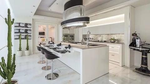 Modern white kitchen with a large island, black bar stools, pendant light fixture, and tall cacti in pots.