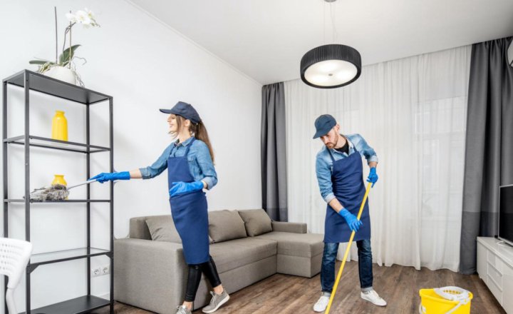 Two professional cleaners in aprons and gloves cleaning a modern living room with a mop and duster.