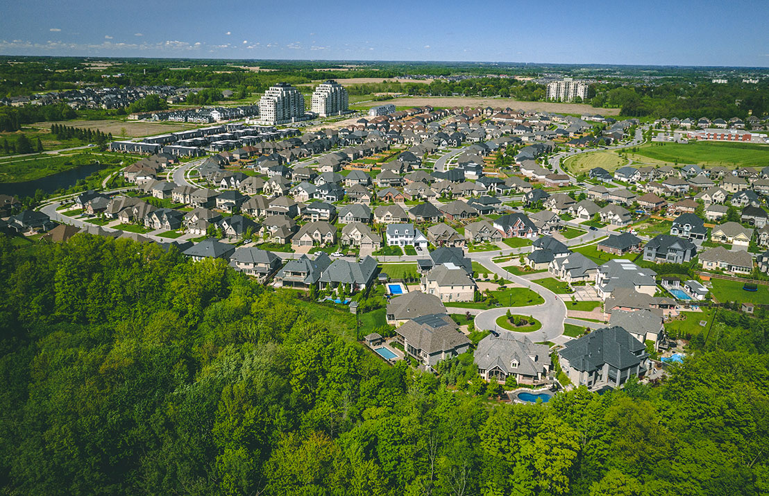 Aerial view of a suburban neighborhood with numerous houses, green lawns, swimming pools, and surrounding trees under a clear blue sky.