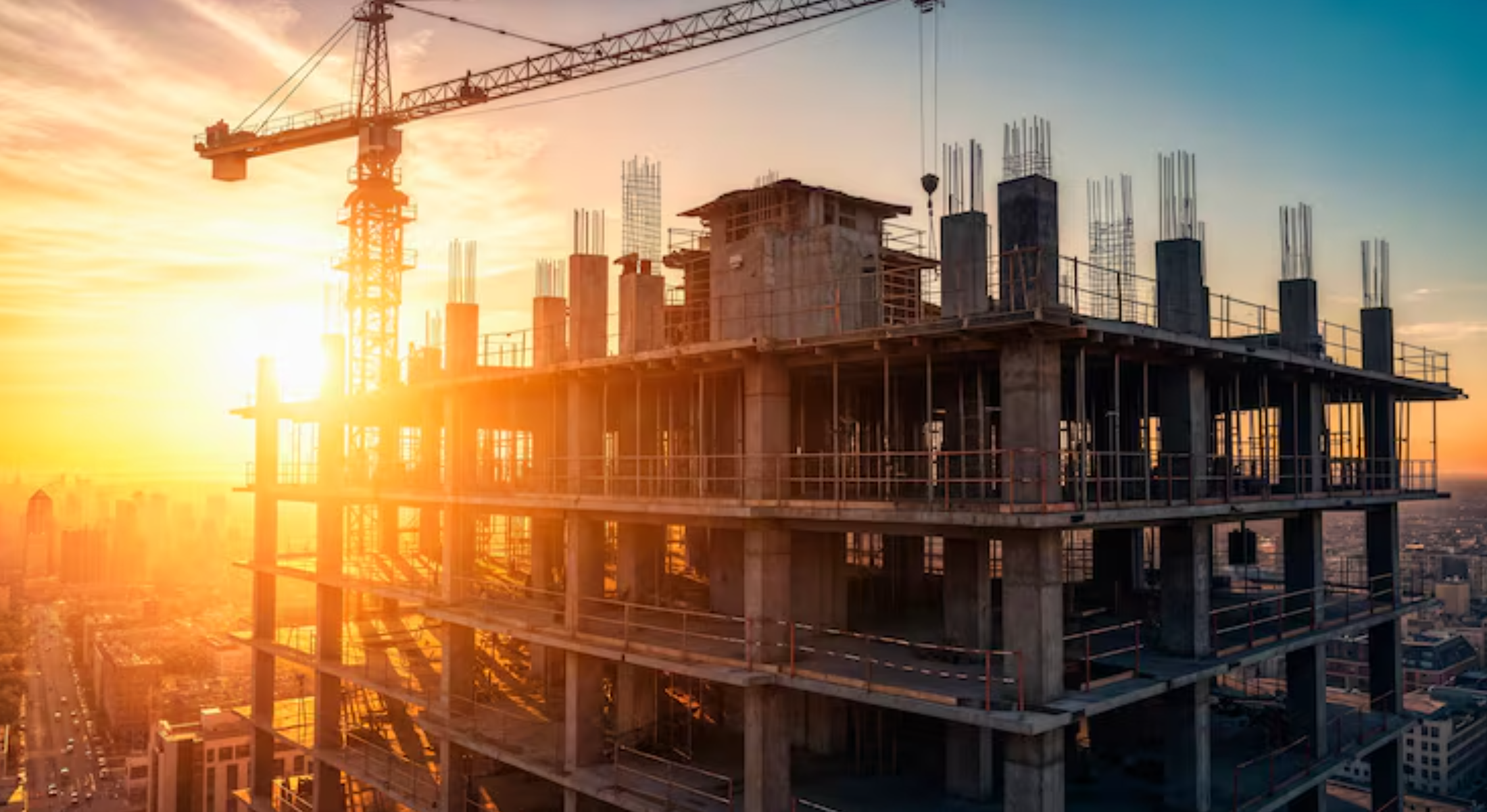 High-rise building under construction with concrete framework and a crane, backlit by the setting sun over a cityscape.