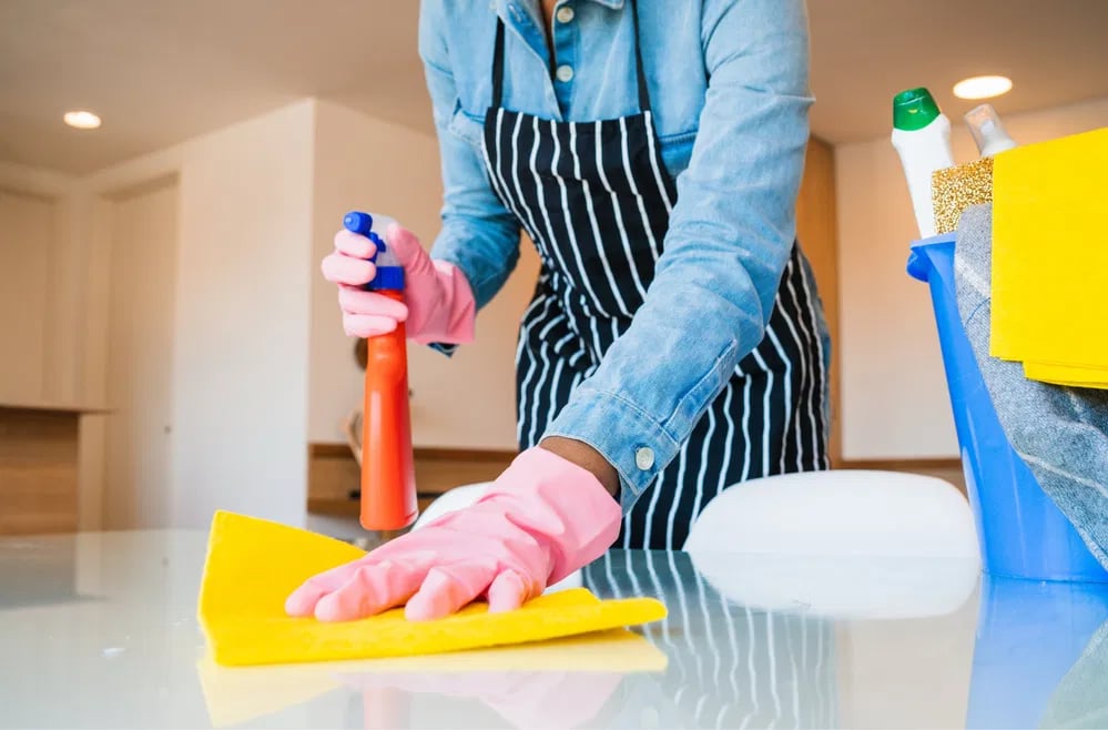 Person wearing pink gloves and a striped apron cleaning a glass table with a yellow cloth and spray bottle.