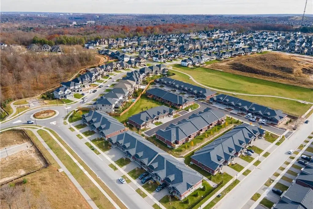 Aerial view of a suburban neighborhood with rows of houses, lawns, and a roundabout surrounded by trees and open land.